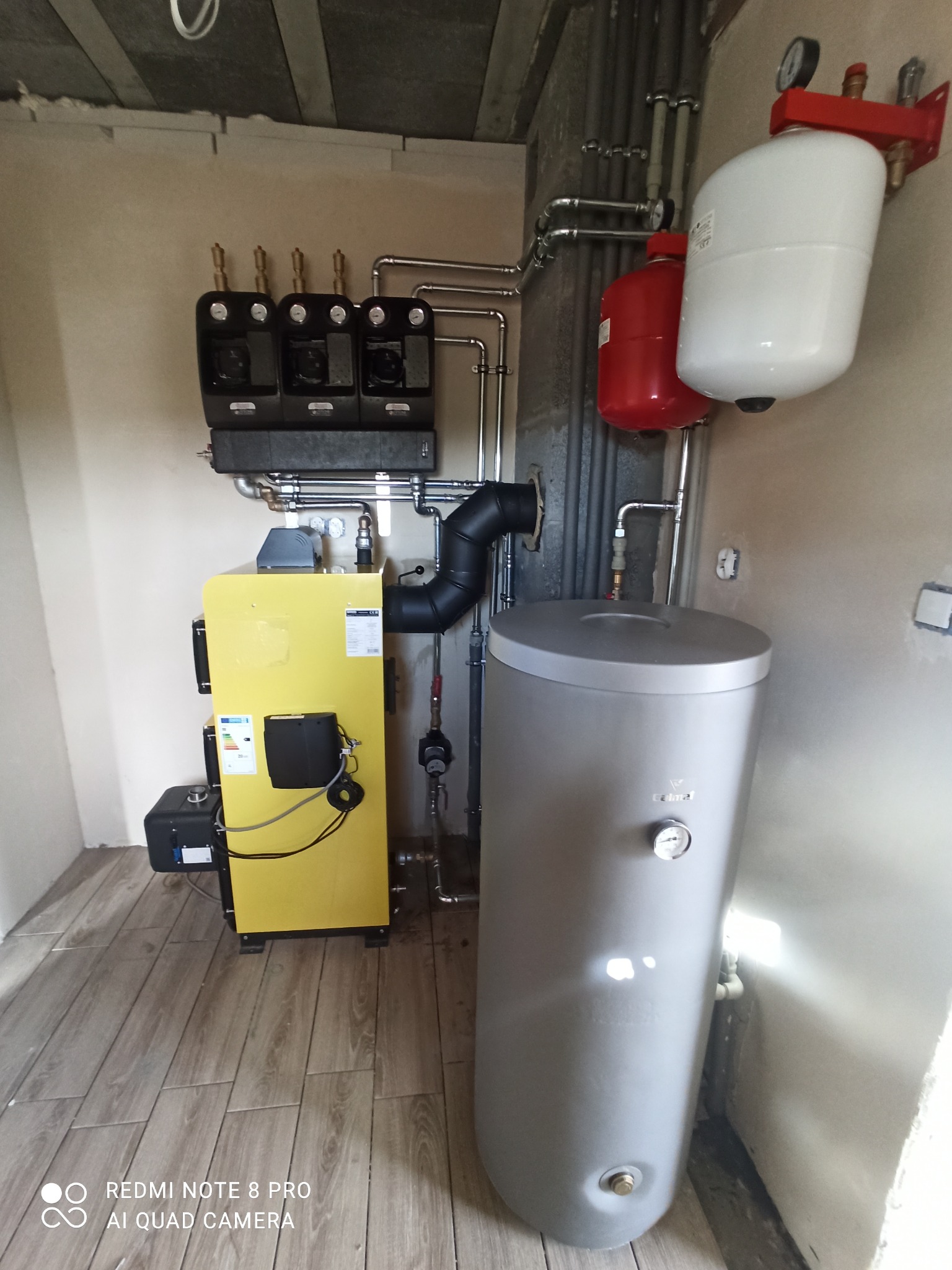 Żółty piec grzewczy z czarnymi elementami, szary zbiornik na wodę, czerwony i biały zbiornik wyrównawczy oraz instalacja rurowa w pomieszczeniu z wykończoną podłogą.