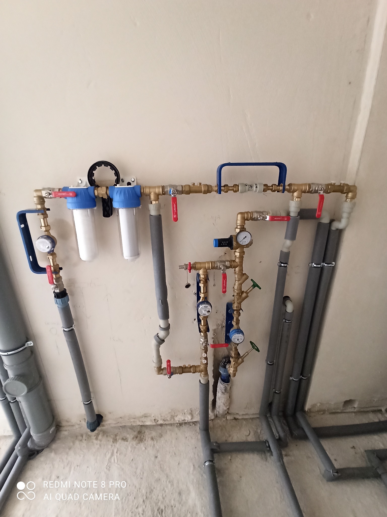 Kompleksowa instalacja wodna z filtrami, manometrami i zaworami na ścianie, widoczne rury szare izolowane i mosiężne elementy.