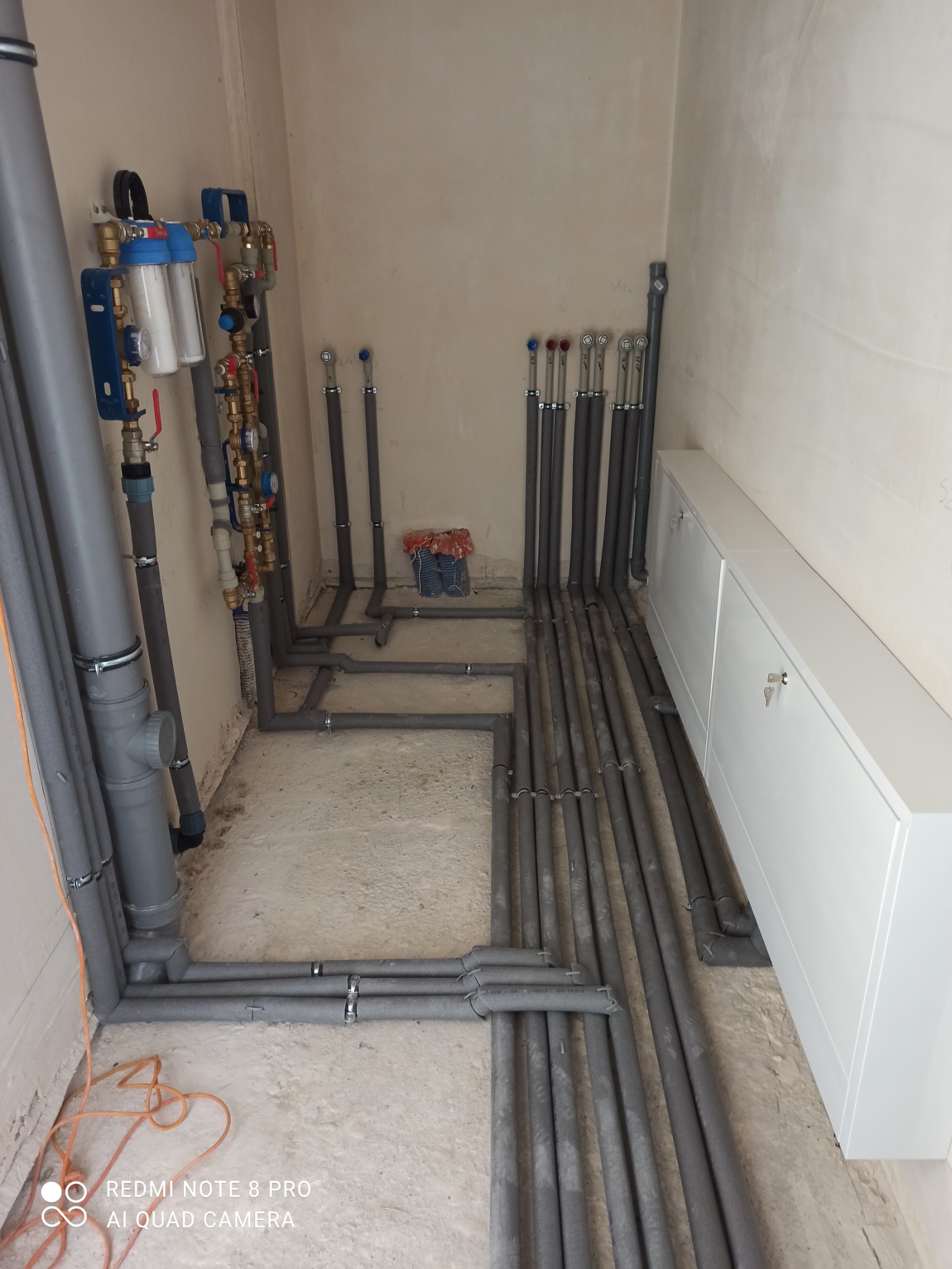 Szare rury instalacyjne w izolacji termicznej ułożone na podłodze w surowym pomieszczeniu, widoczne filtry wody, biała szafka w tle.