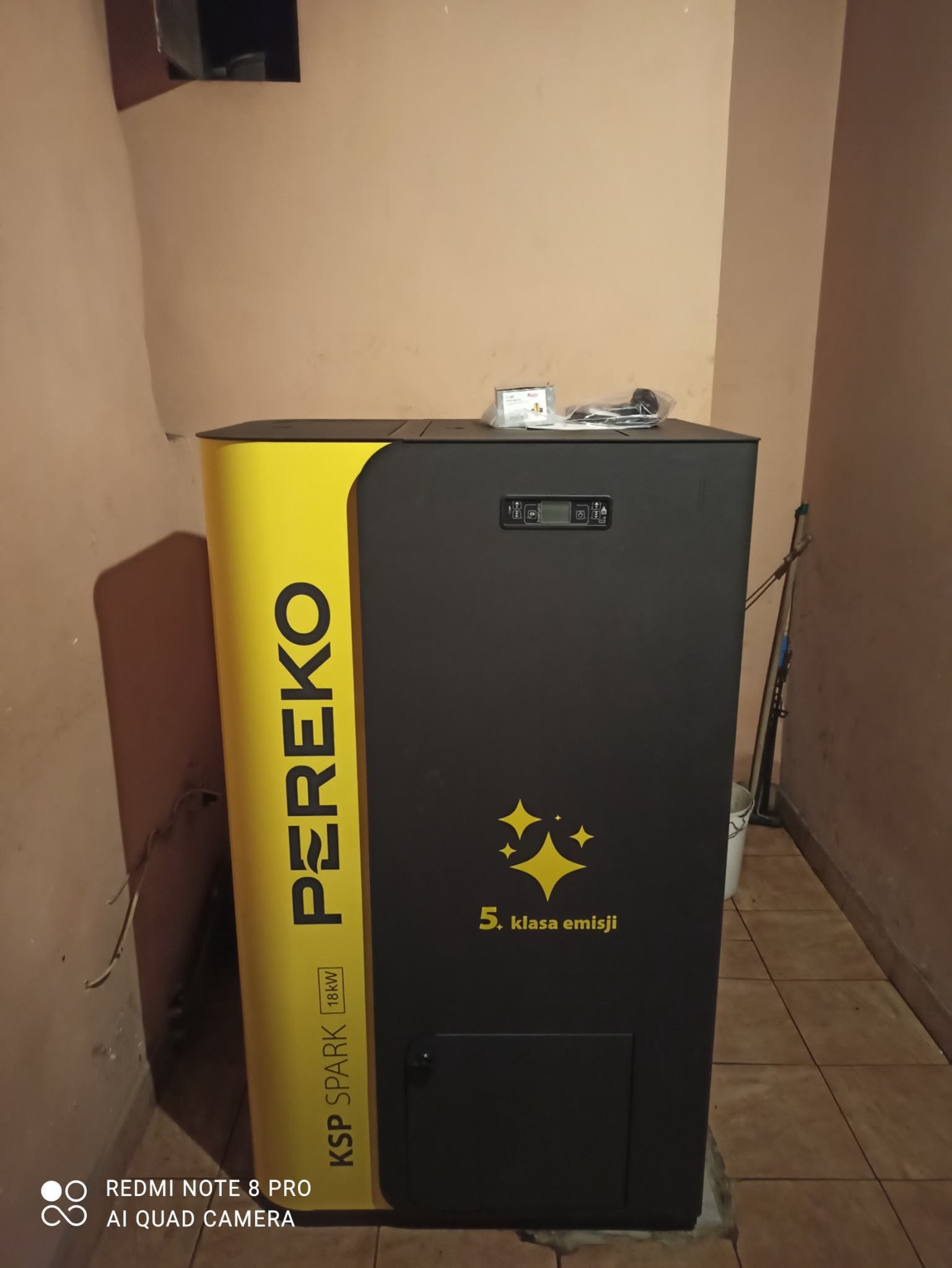 Kocioł grzewczy marki PEREKO KSP SPARK 18kW, oznaczony jako 5. klasa emisji, stojący w rogu pomieszczenia z widocznymi akcesoriami na górze.