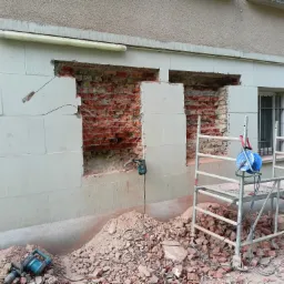 Fragment ściany budynku z dwoma wyburzonymi otworami, odsłaniającymi czerwoną cegłę, z gruzem u podstawy i rusztowaniem obok, widoczny młot pneumatyczny.