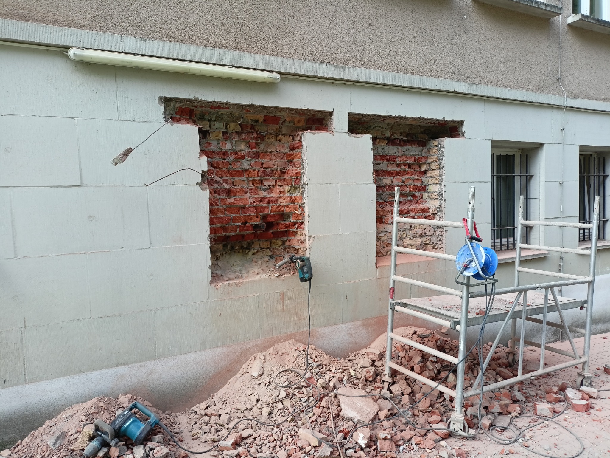 Fragment ściany budynku z dwoma wyburzonymi otworami, odsłaniającymi czerwoną cegłę, z gruzem u podstawy i rusztowaniem obok, widoczny młot pneumatyczny.