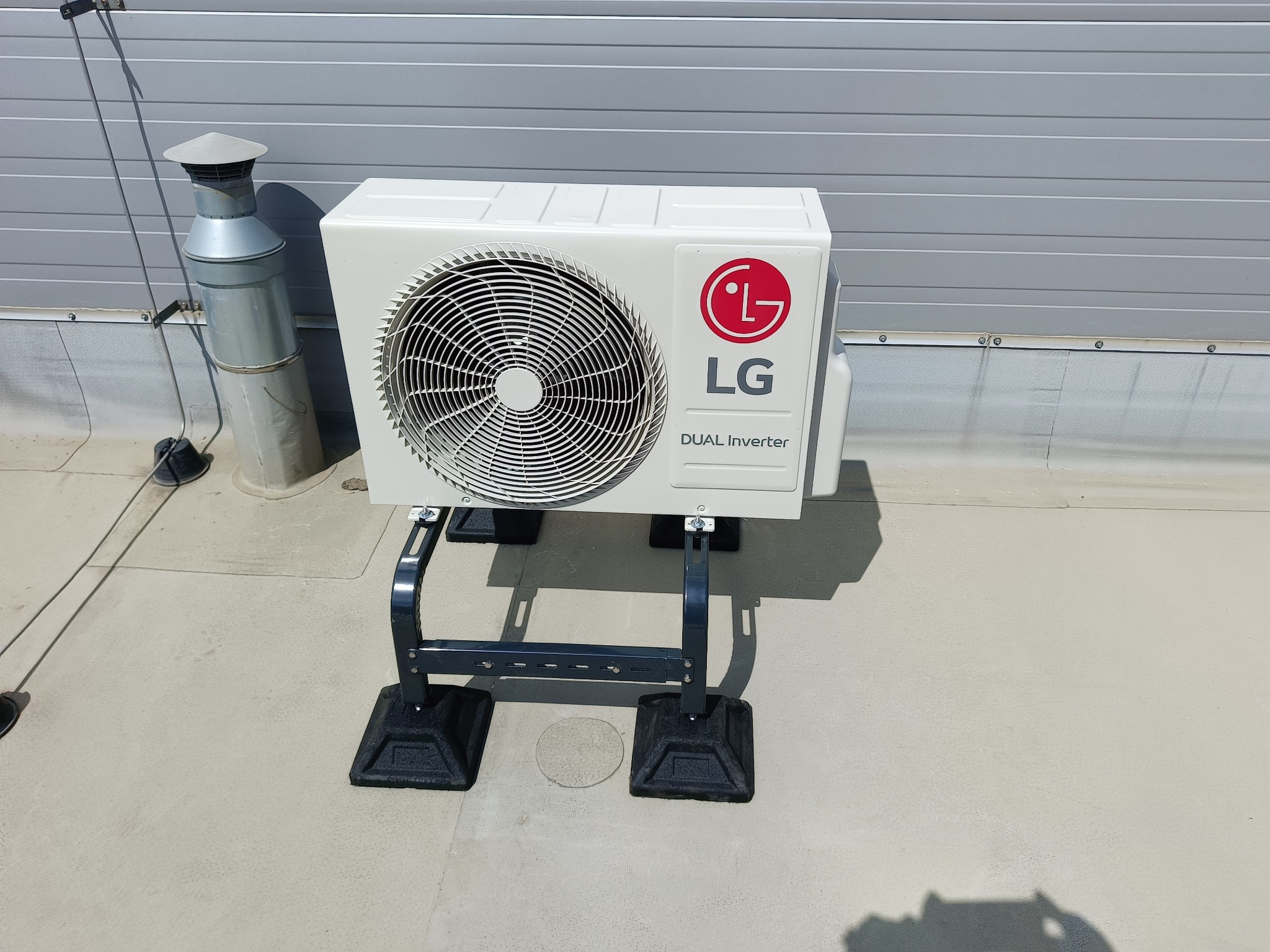 Zewnętrzna jednostka klimatyzacji LG Dual Inverter zamontowana na dachu, widoczny komin wentylacyjny w tle.