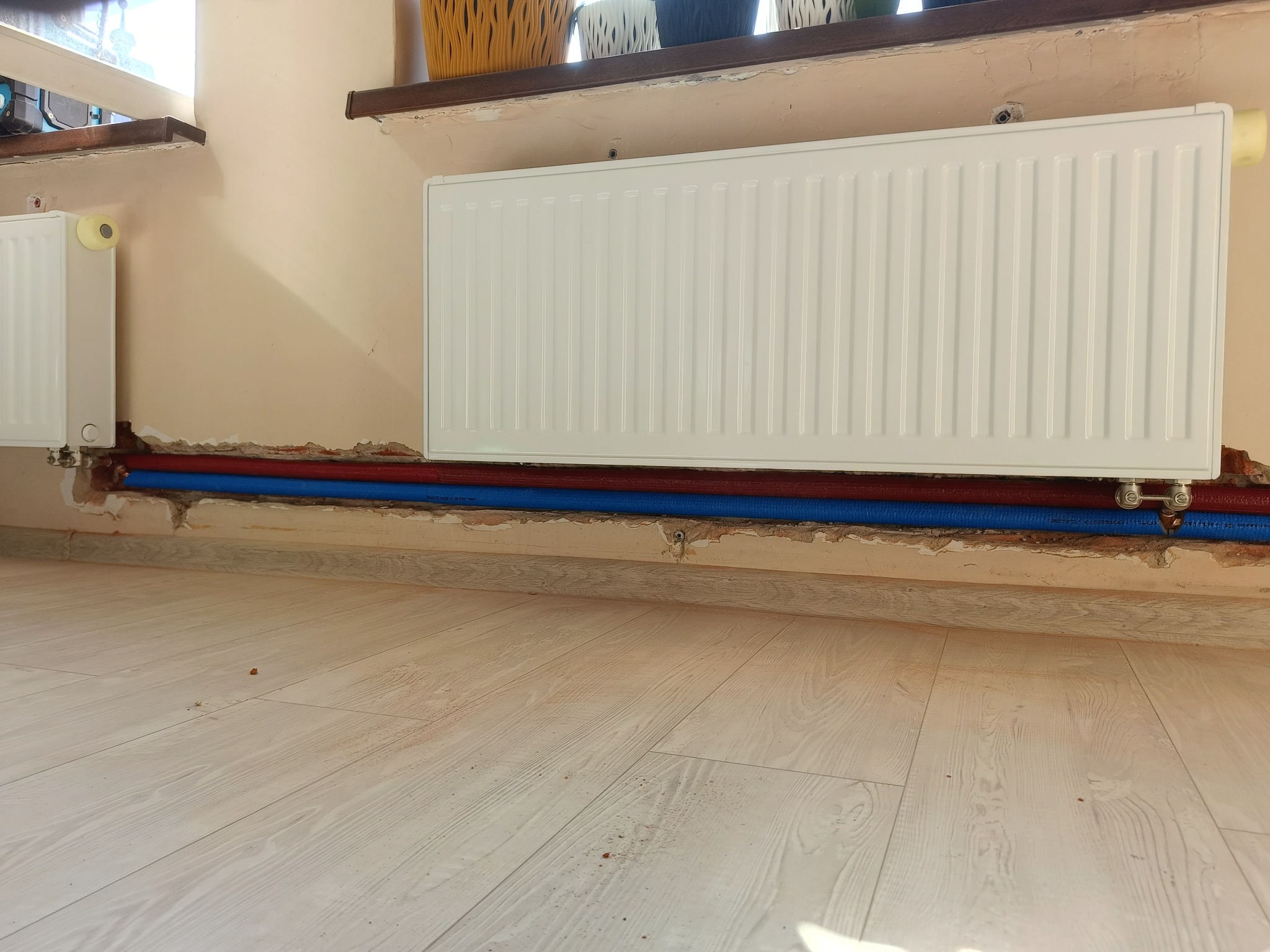 Biały grzejnik panelowy zamontowany na ścianie, widoczne czerwone i niebieskie rury instalacji grzewczej, ślady po pracach remontowych na ścianie i podłodze.