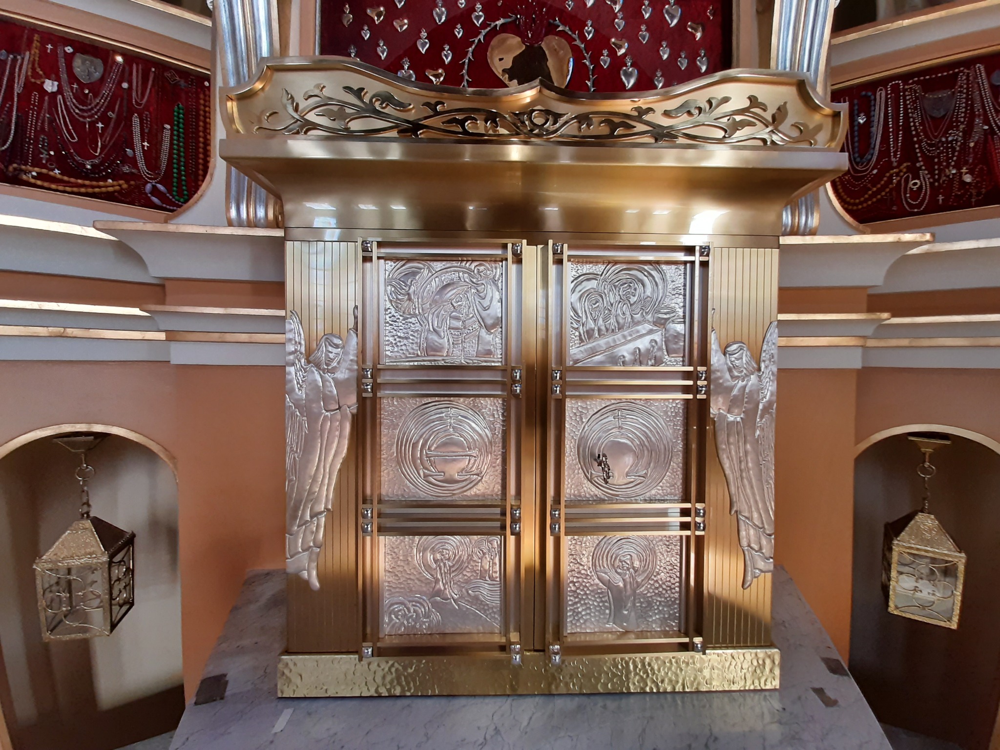 Zdobiona, metalowa tabernakulum z płaskorzeźbami aniołów i scen religijnych, umieszczona w kościele, oświetlona ciepłym światłem.