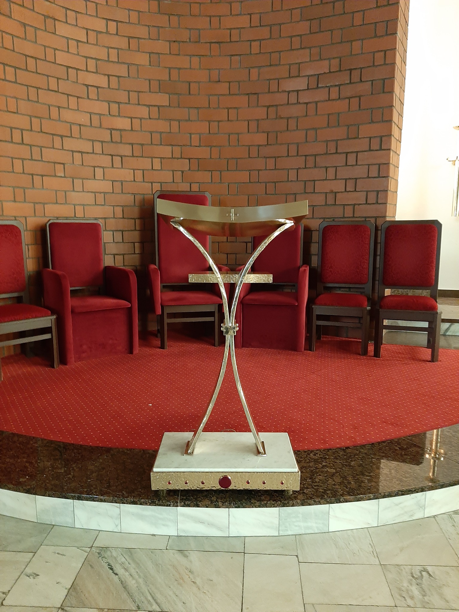 Metalowy pulpit z marmurową podstawą na tle ceglanej ściany i czerwonych krzeseł z aksamitną tapicerką.