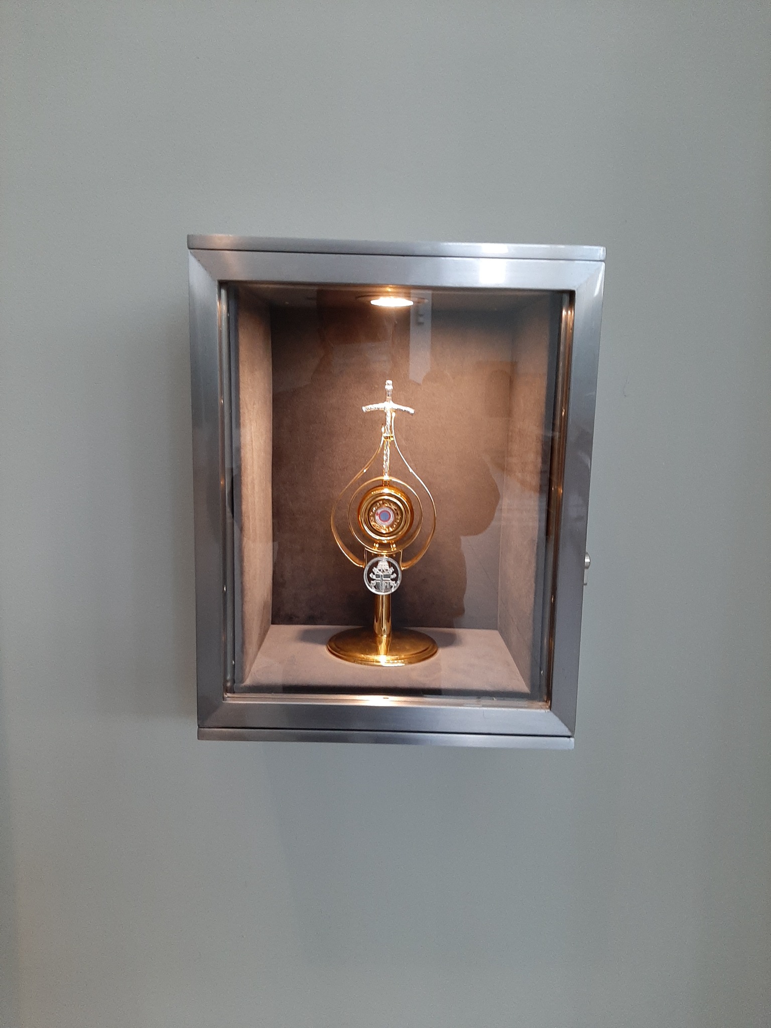 Złota monstrancja z krzyżem i fioletowym kamieniem, umieszczona w przeszklonej, metalowej gablocie z wewnętrznym oświetleniem, zamontowanej na szarej ścianie.