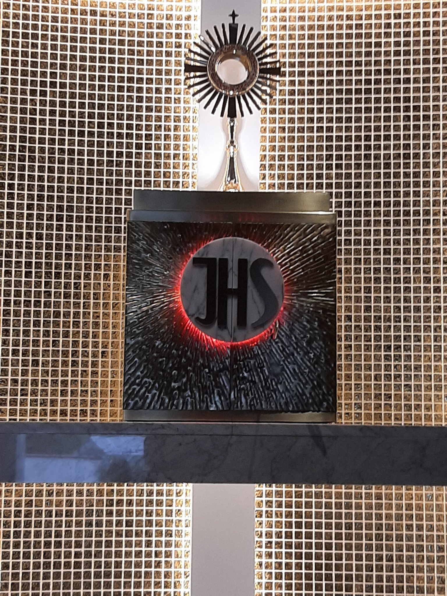 Monstrancja z symbolem IHS na tle ściany z mozaiki w złotych odcieniach.