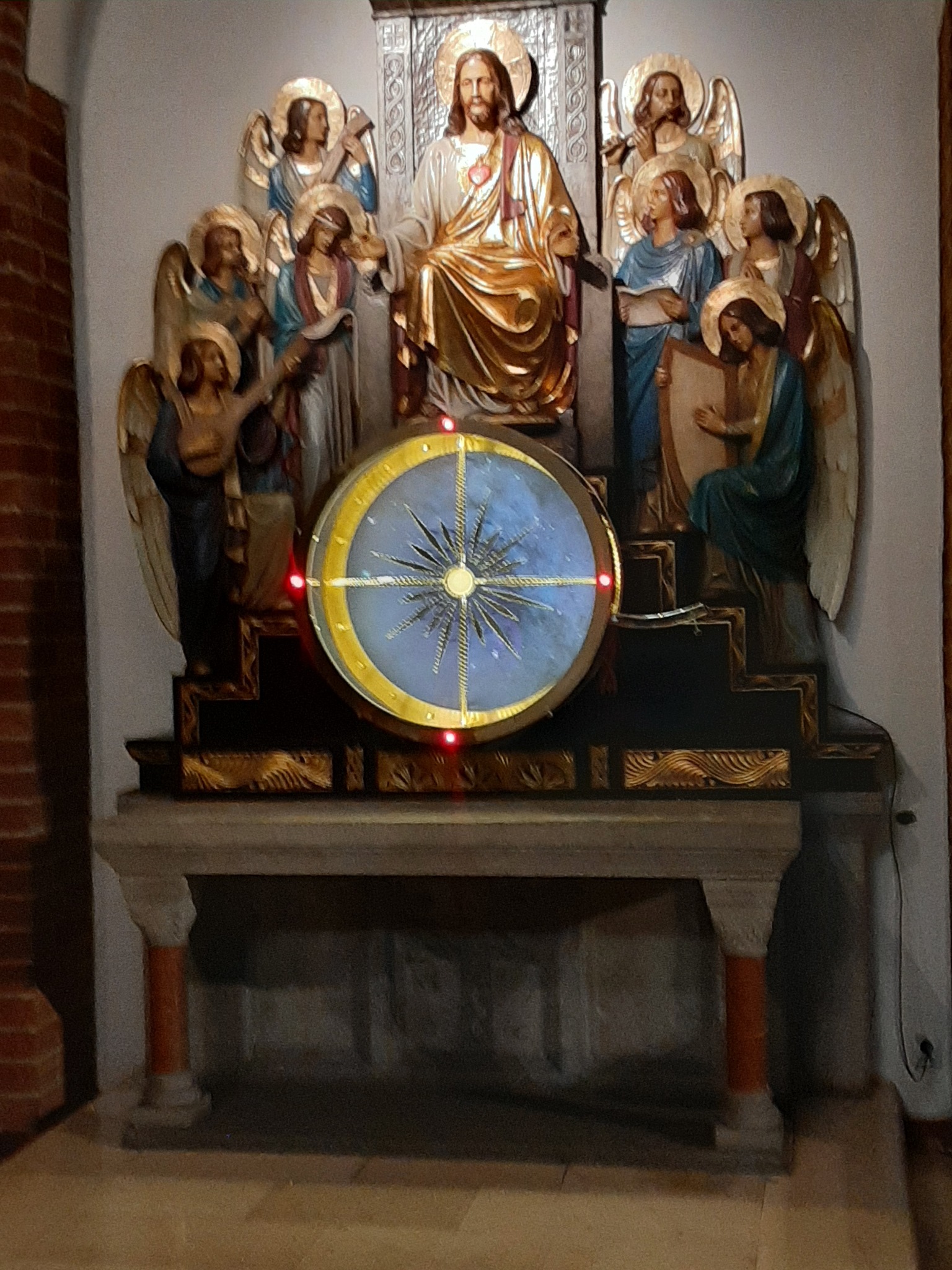 Rzeźbiona kompozycja religijna z Jezusem na tronie, otoczonym przez anioły, z podświetlanym, okrągłym elementem dekoracyjnym z motywem gwiazdy na pierwszym planie, umieszczona na kamiennym...