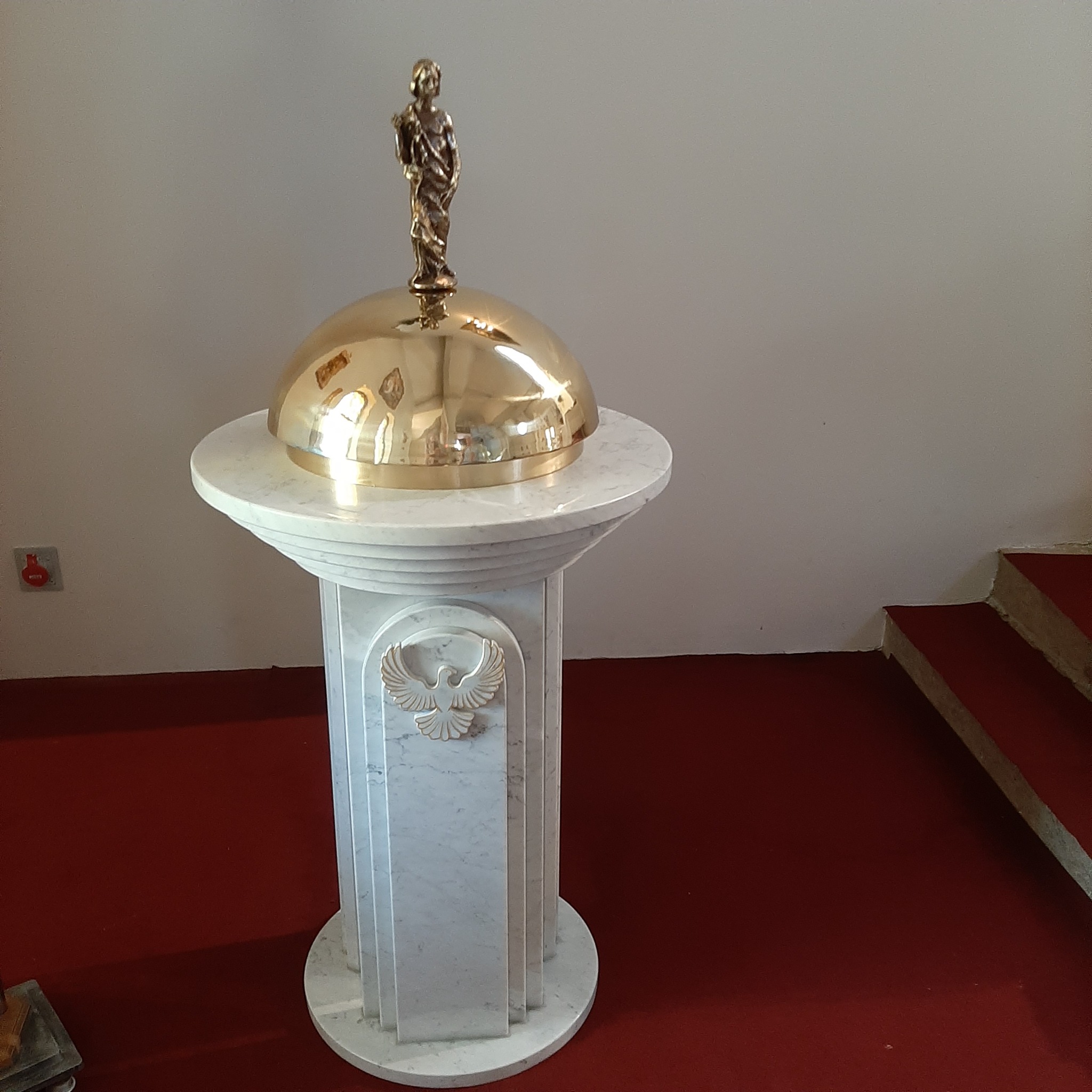 Marmurowa chrzcielnica z pozłacaną kopułą i figurką, umieszczona na czerwonej posadzce w kościele.