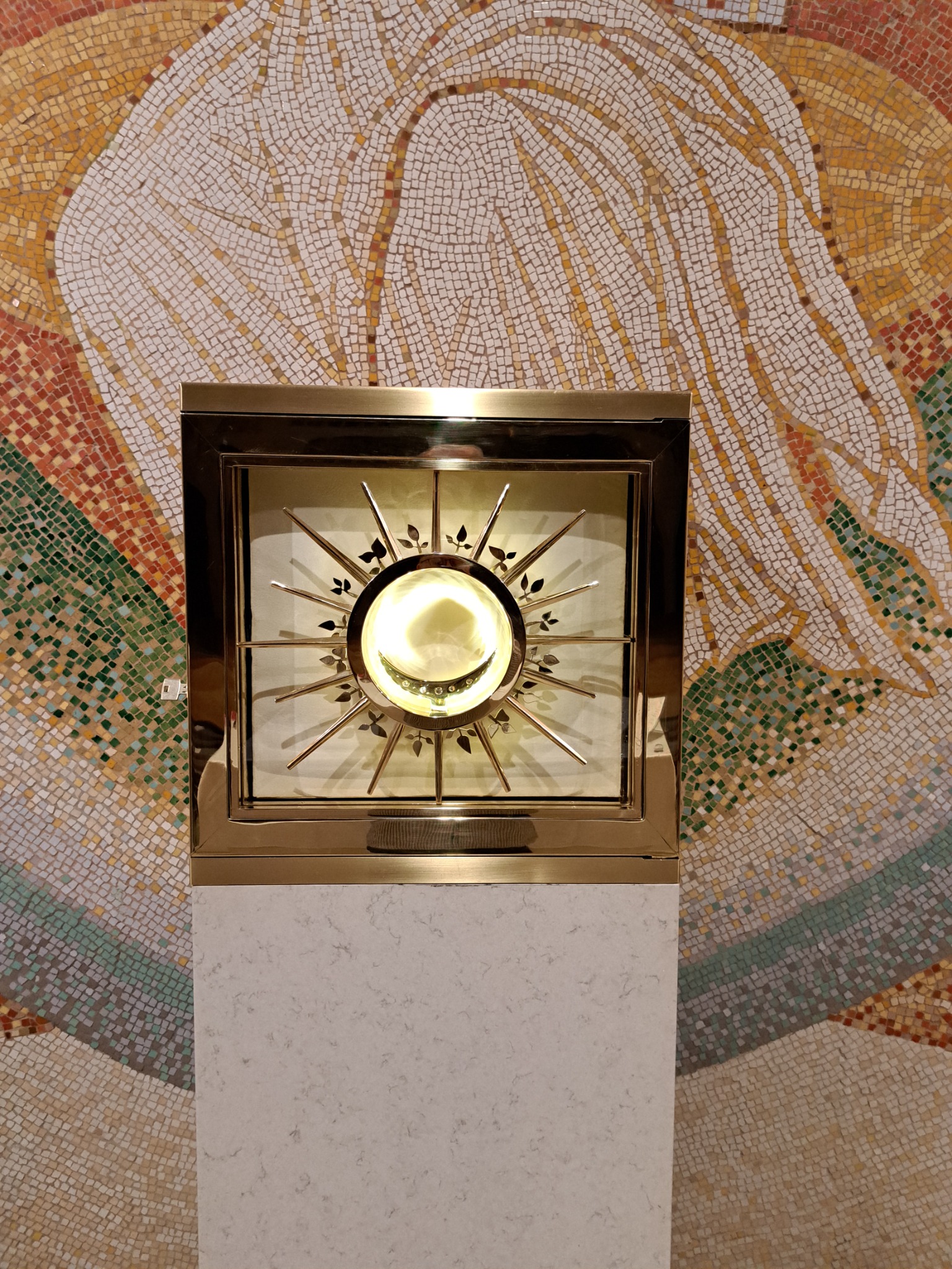 Złota, kwadratowa gablota z promieniście ułożonymi metalowymi prętami i dekoracjami w kształcie liści, umieszczona na białym postumencie, na tle mozaiki z drobnych kwadratowych elementów...