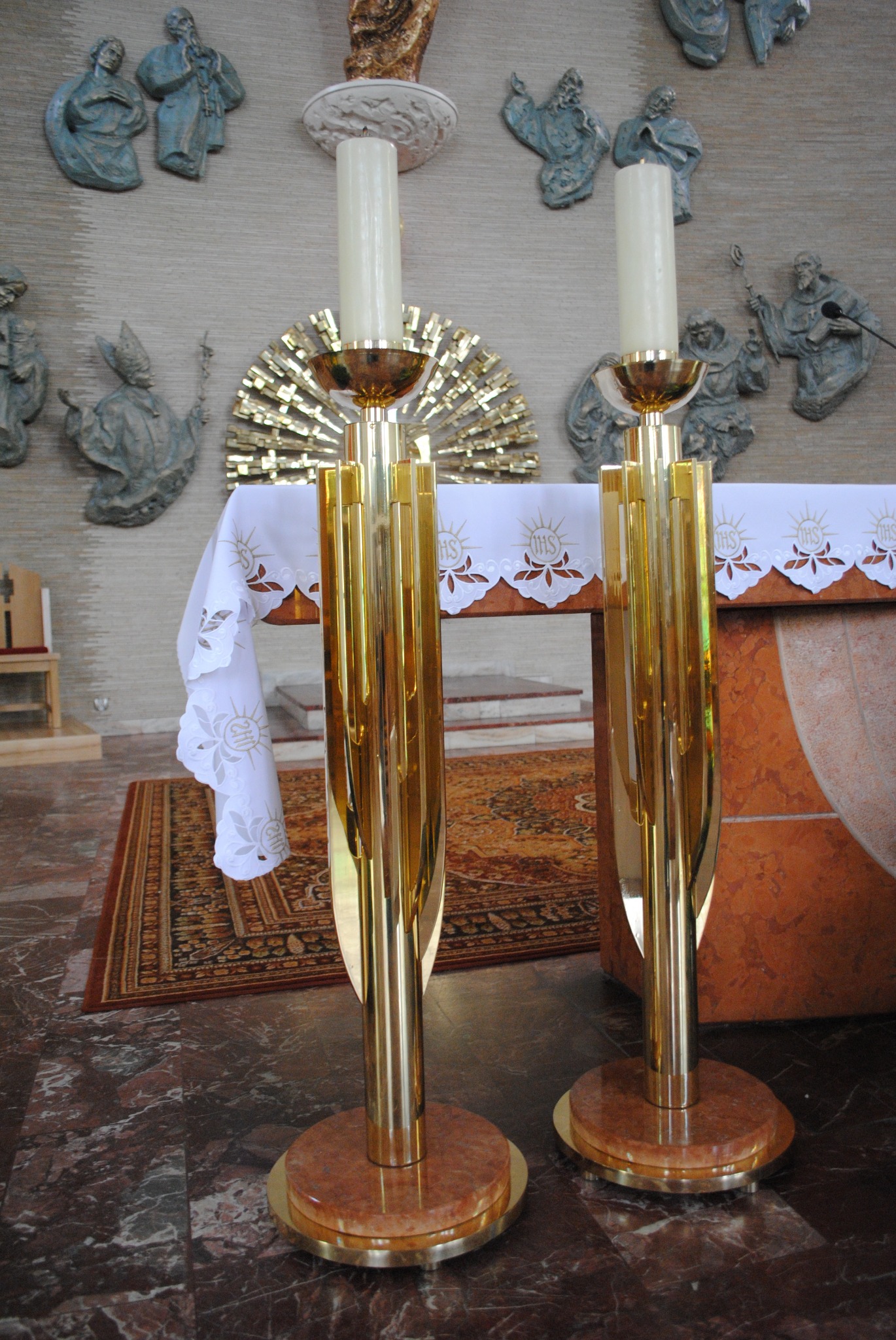 Dwa złote, metalowe świeczniki z zapalonymi świecami na marmurowej posadzce, z ołtarzem i płaskorzeźbami na ścianie w tle.