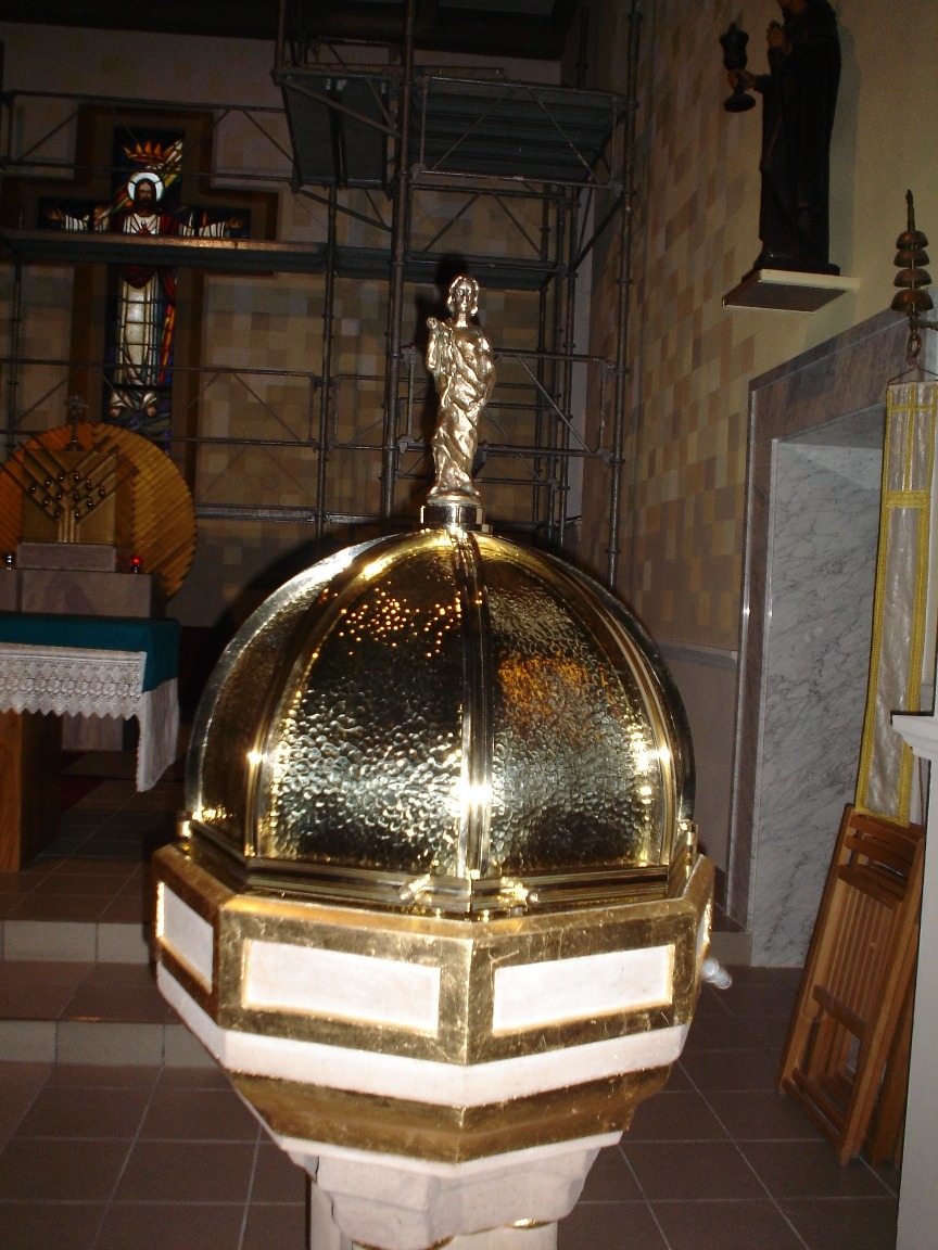 Złota chrzcielnica z figurką na kopule, widoczna w kościele podczas remontu, z rusztowaniami w tle.