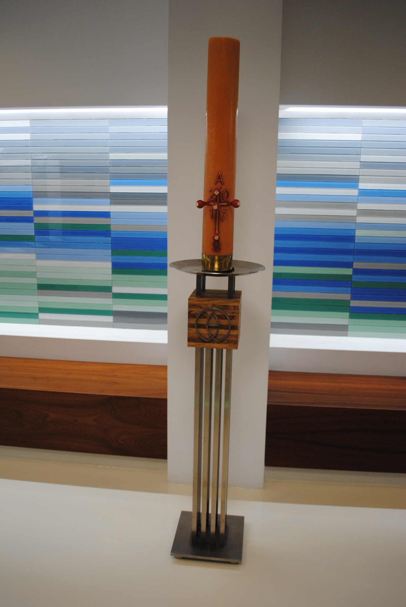 Metalowy świecznik z drewnianą podstawą i brązową świecą z datą 2017, na tle ściany z kolorowymi pasami.