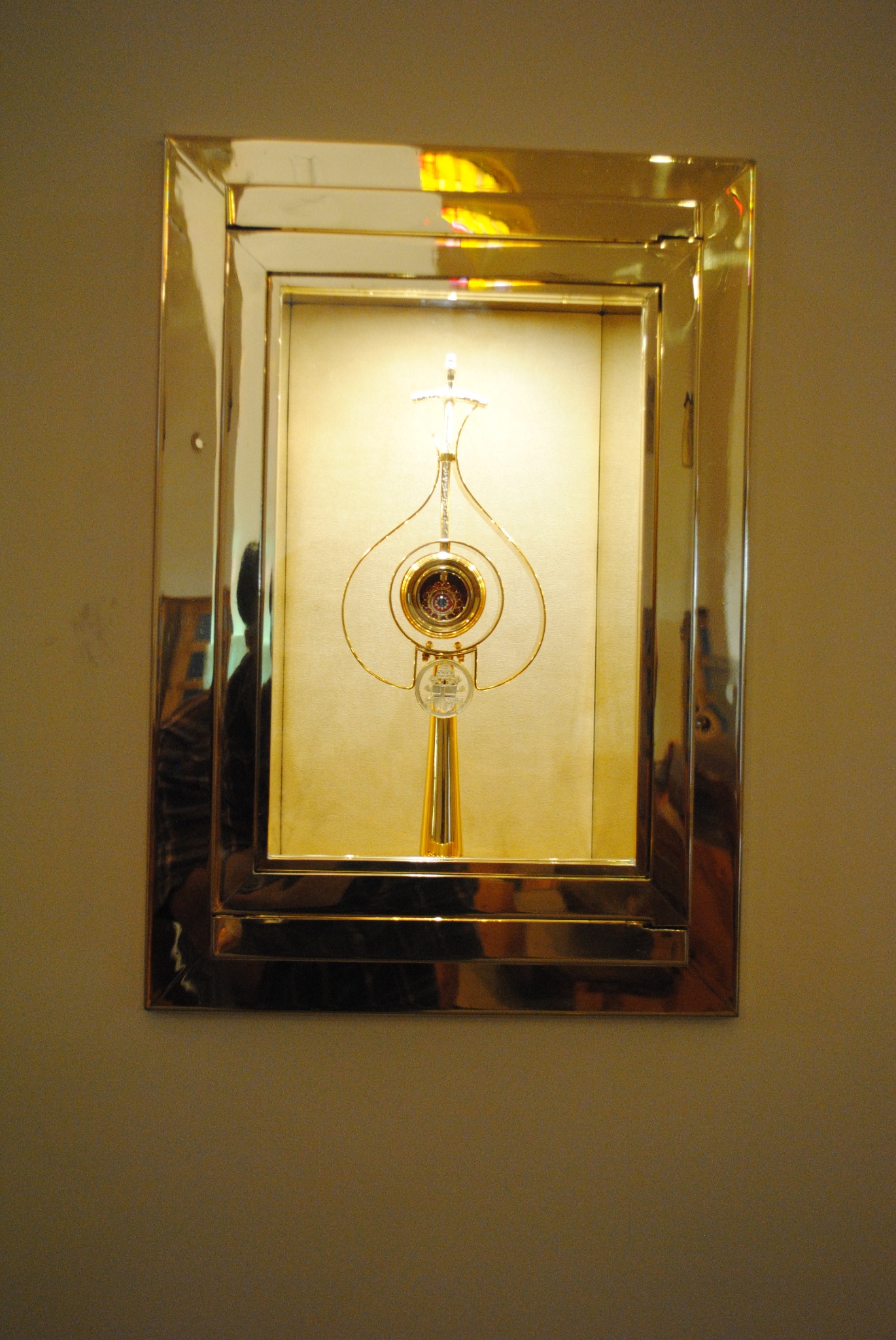 Złota, ozdobna relikwia w szklanej gablocie z polerowanej stali, zawieszona na ścianie.