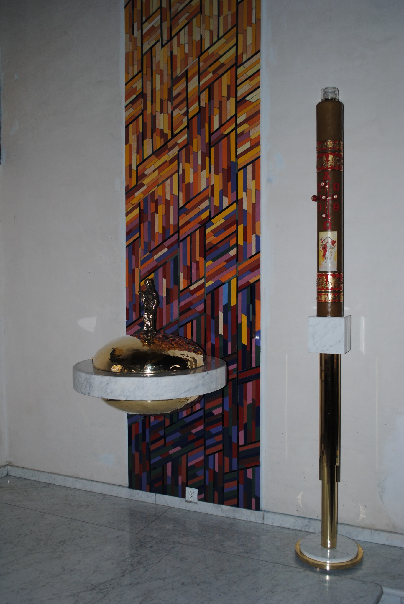 Złota misa z figurką na marmurowym postumencie, obok wysoka świeca na złotym stojaku z marmurową podstawą, na tle ściany z geometrycznym wzorem z kolorowych prostokątów.