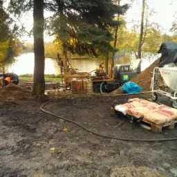 Wiertnica do prac ziemnych na błotnistym terenie nad jeziorem, widoczny żółty traktor, paleta z workami cementu i wózek z pojemnikiem, w tle drzewa w jesiennych barwach.