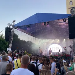 Scena plenerowa z muzykami podczas koncertu, nagłośnienie po bokach, tłum ludzi przed sceną, w tle budynek z wieżą.