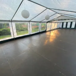 Wnętrze pustego namiotu imprezowego z podłogą z ciemnych płyt, oświetlonego wiszącymi papierowymi kulami i promieniami popołudniowego słońca wpadającymi przez okna.