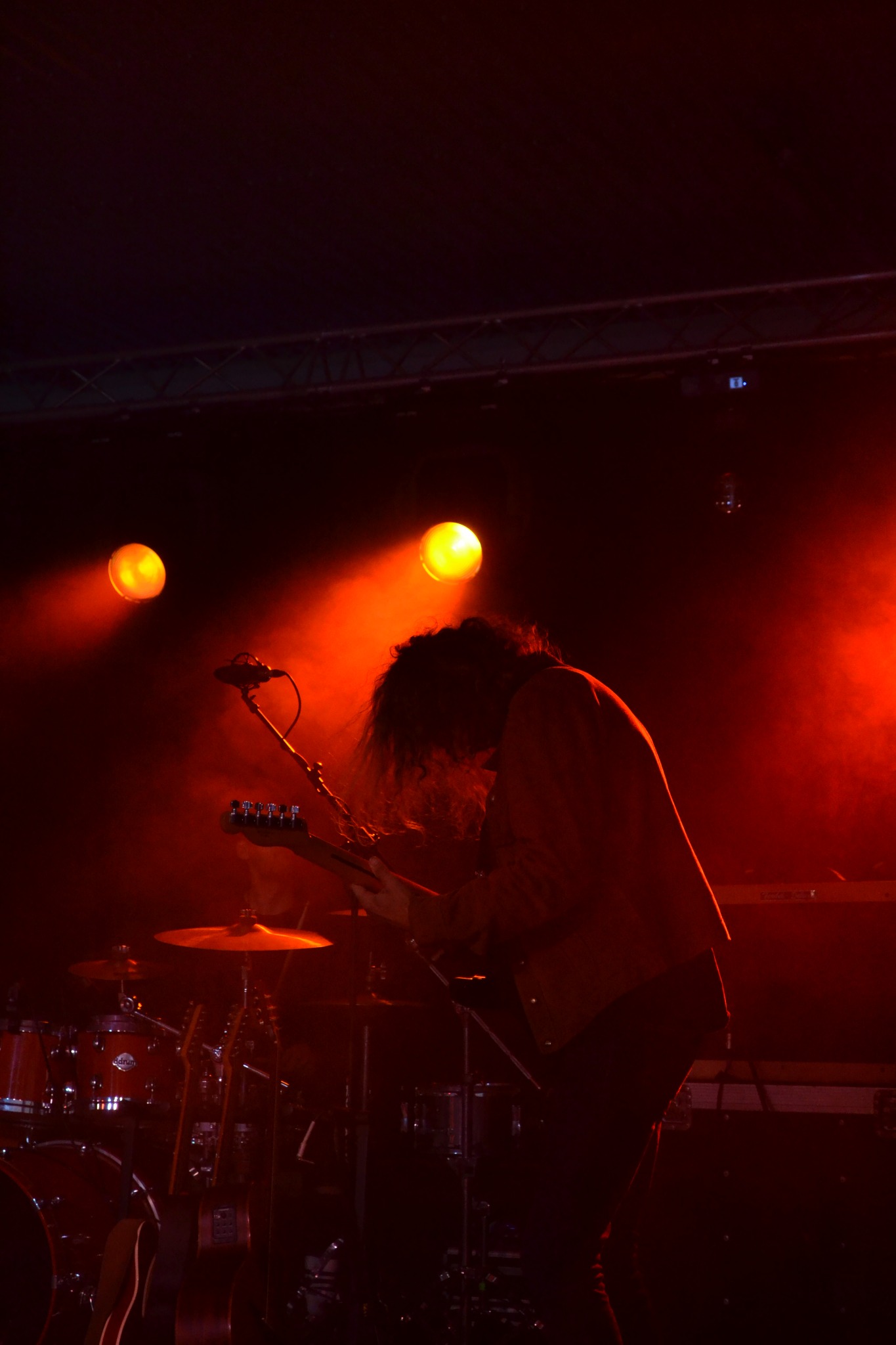 Gitarzysta w czerwonym oświetleniu gra na gitarze podczas koncertu, widoczna perkusja i elementy sceny w tle.