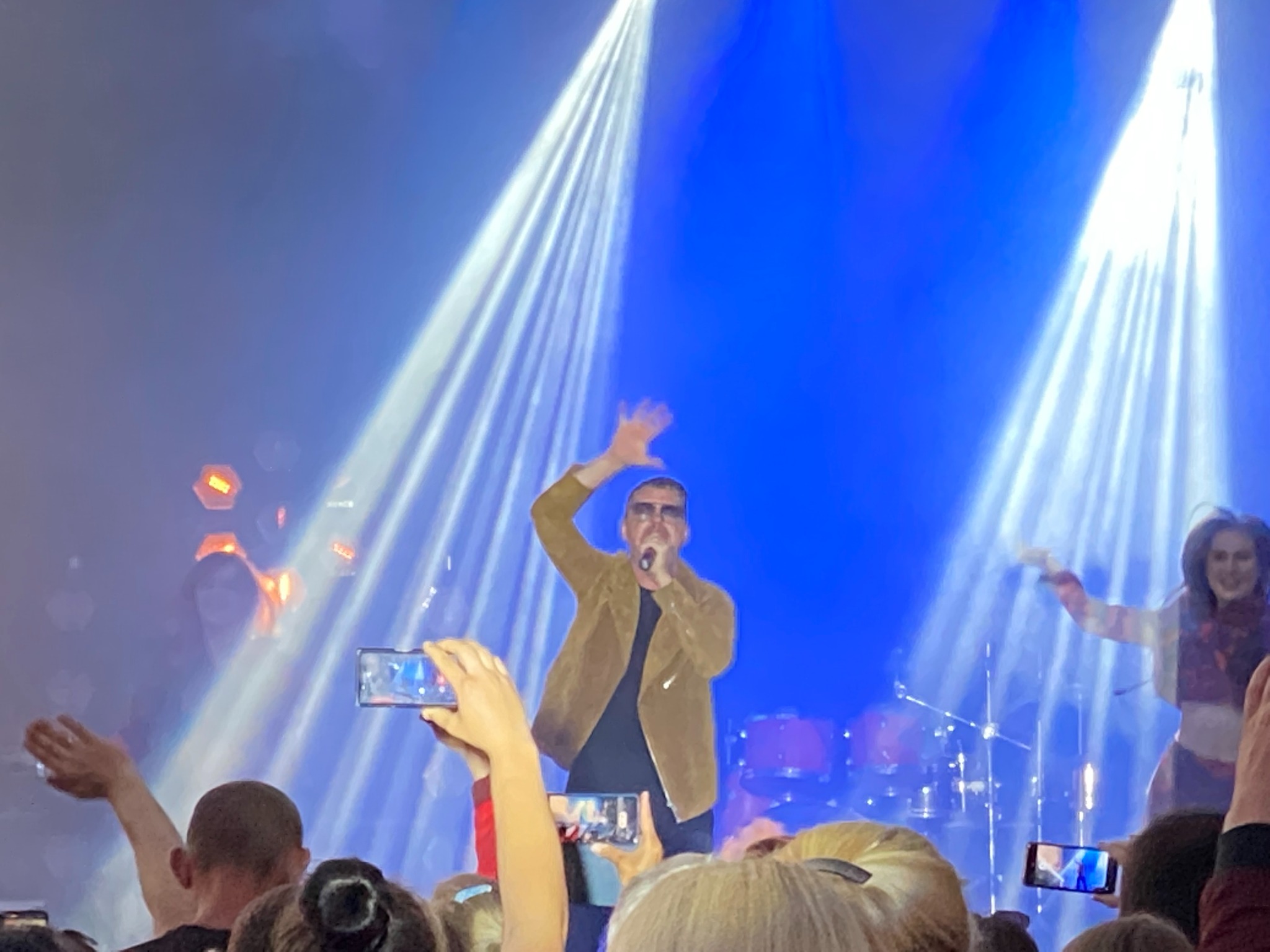Wokalista w brązowej kurtce śpiewa do mikrofonu na scenie oświetlonej białymi reflektorami, widzowie filmują występ telefonami.