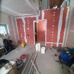 Wnętrze w trakcie montażu płyt kartonowo-gipsowych na ścianach i suficie, widoczne narzędzia, worki z zaprawą, puszki z farbą oraz wystające kable elektryczne.