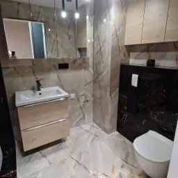 Nowoczesna łazienka z płytkami imitującymi marmur, wisząca szafka z umywalką, czarna zabudowa toalety i podwieszane lampy punktowe.