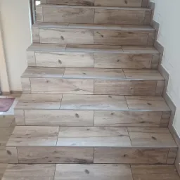 Schody wyłożone płytkami imitującymi drewno, z widocznymi fugami i listwami przypodłogowymi, częściowo wykończone, z widocznymi śladami prac wykończeniowych na ścianach.