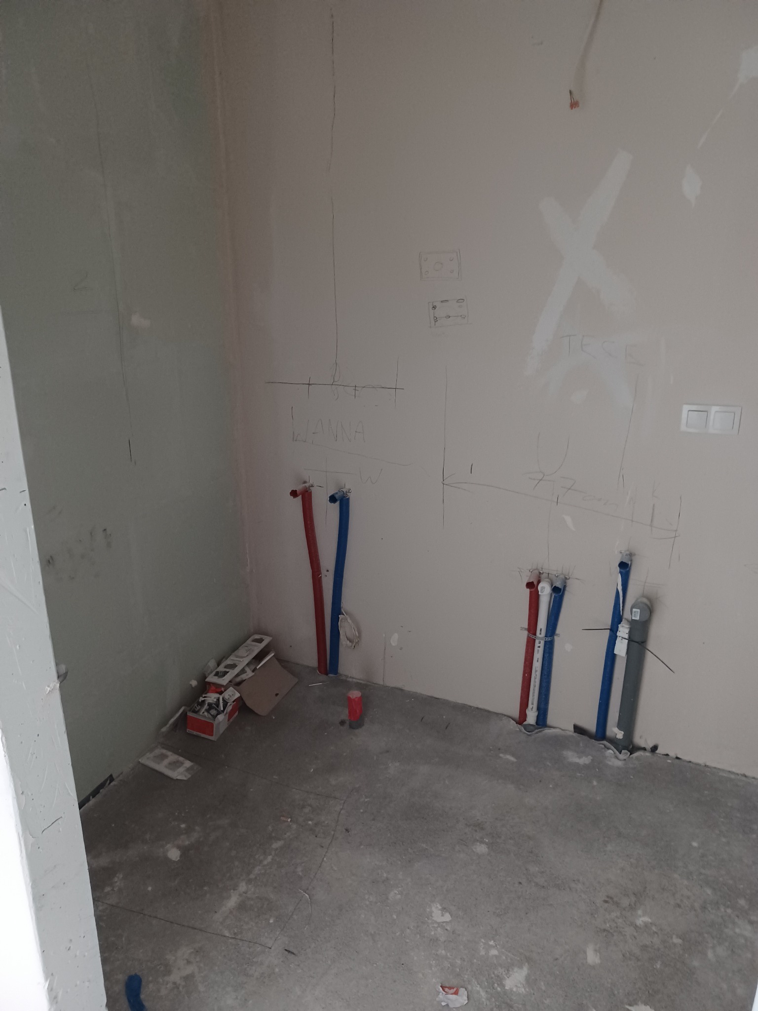 Surowe wnętrze łazienki w trakcie prac instalacyjnych: widoczne wyprowadzenia rur wodnych oznaczonych kolorami, szkic na ścianie z wymiarami, puszki elektryczne, podłoże betonowe.