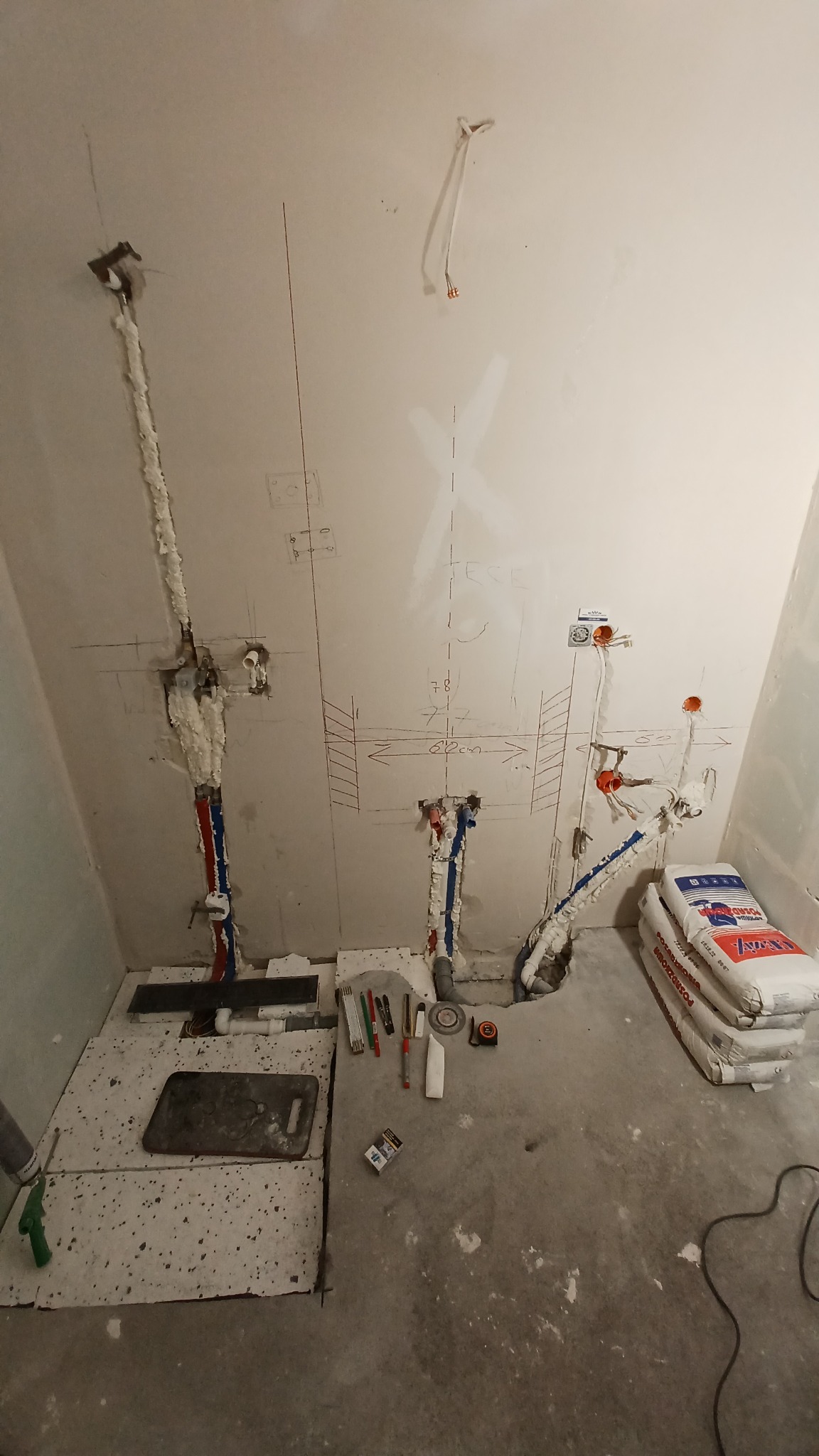 Łazienka w trakcie remontu: widoczne rury wodne i kanalizacyjne, częściowo zaizolowane pianką montażową, zaznaczone na ścianie punkty instalacji, worki z zaprawą, narzędzia glazurnicze i fragment...