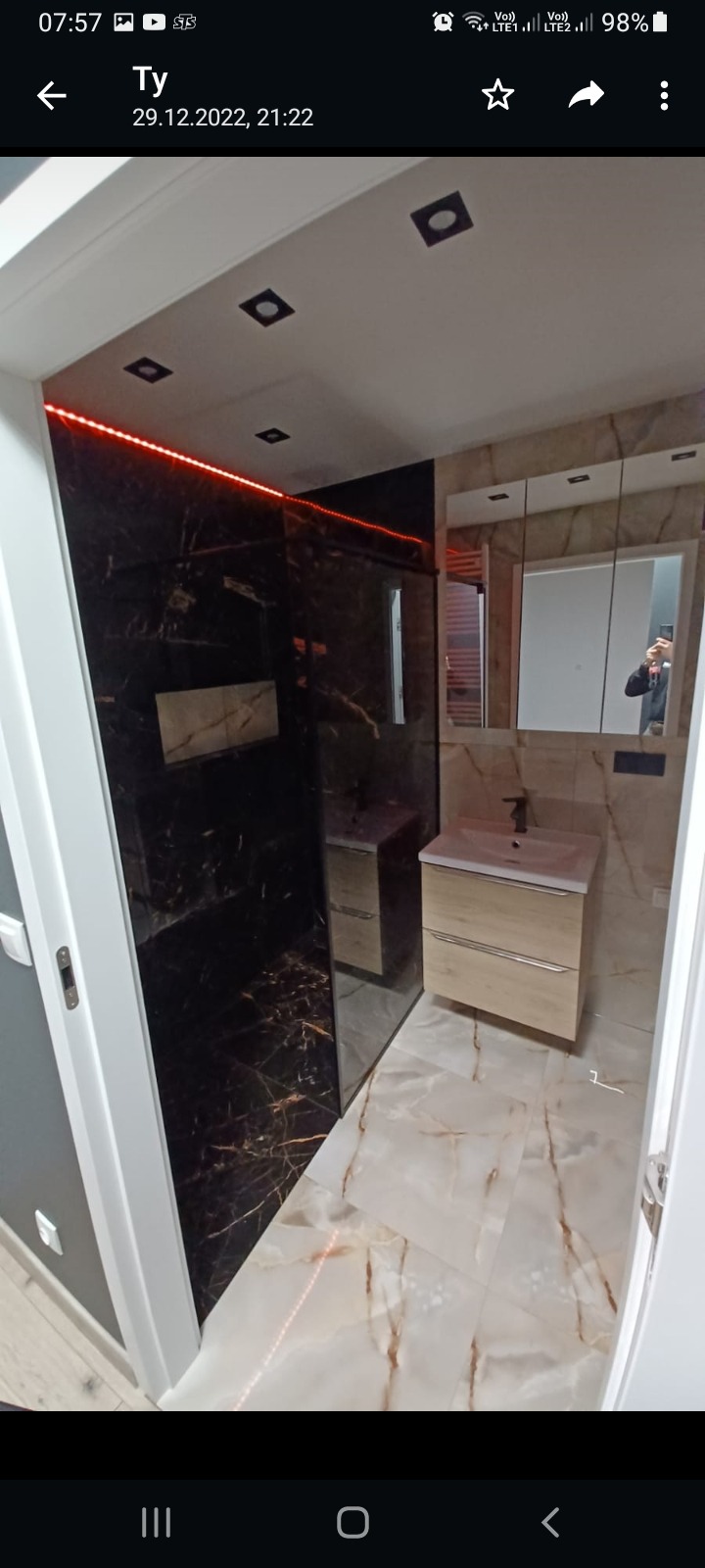 Nowoczesna łazienka z czarną kabiną prysznicową i jasną umywalką z szafką, podświetlenie LED wzdłuż sufitu i podłogi, marmurowe płytki na podłodze i ścianach.