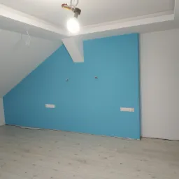 Pokój na poddaszu po remoncie: ściany pomalowane na biało i turkusowo, jasne panele podłogowe, widoczne gniazdka elektryczne i żarówka na kablu.
