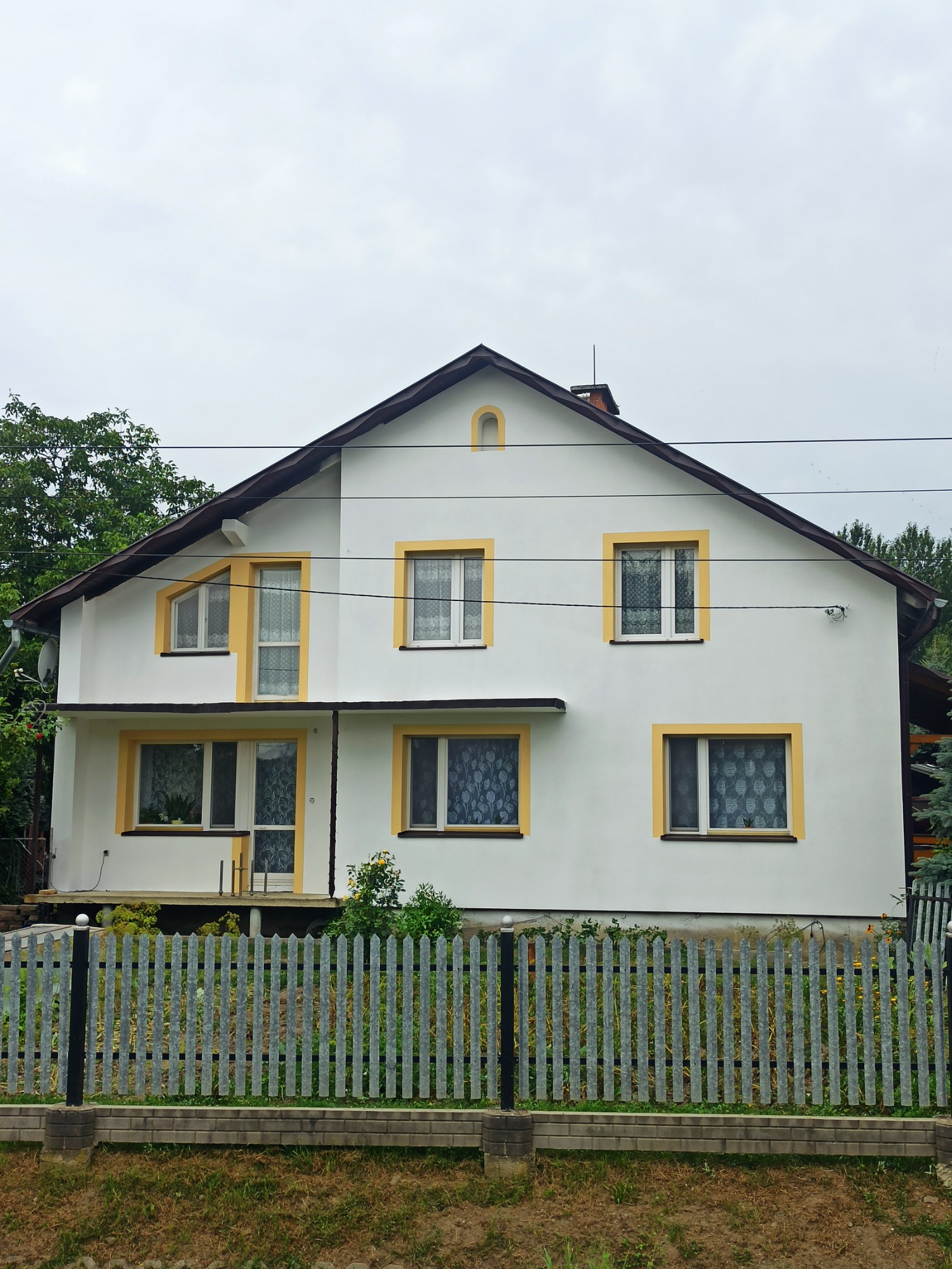 Odnowiona elewacja dwupiętrowego domu z białymi ścianami i żółtymi obramowaniami okien, widoczna spod metalowego ogrodzenia.