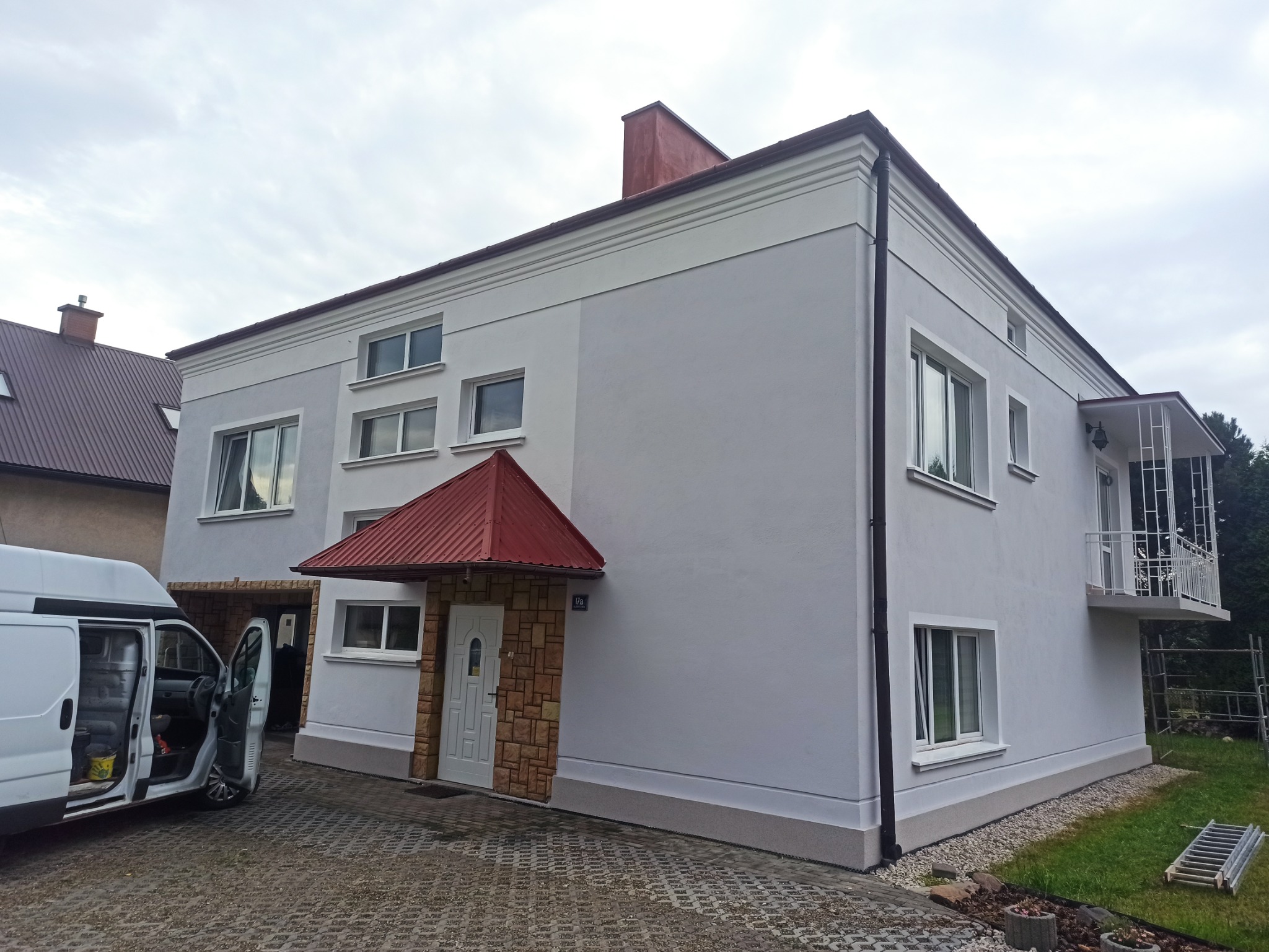 Dwupiętrowy budynek z szarą elewacją, czerwonym daszkiem nad wejściem i białymi ramami okiennymi, widziany z perspektywy lekko z dołu, z otwartym vanem parkującym obok.