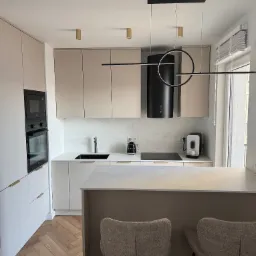 Elegancka kuchnia w beżach i bieli. Nowoczesne oświetlenie, marmurowe blaty, drewniana podłoga w jodełkę. Minimalistyczny design z wyspą i hokerami.