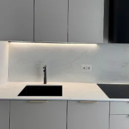 Minimalistyczna kuchnia: szare szafki, czarny zlew i bateria, marmurowy blat z podświetleniem LED. Gniazdka elektryczne na ścianie.