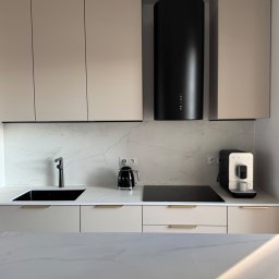 HouseOfDesign - Beżowa kuchnia z czarnymi akcentami: okap cylindryczny, płyta indukcyjna, czajnik i ekspres Smeg. Blat i ściana z białego marmuru. Minimalistyczny design.