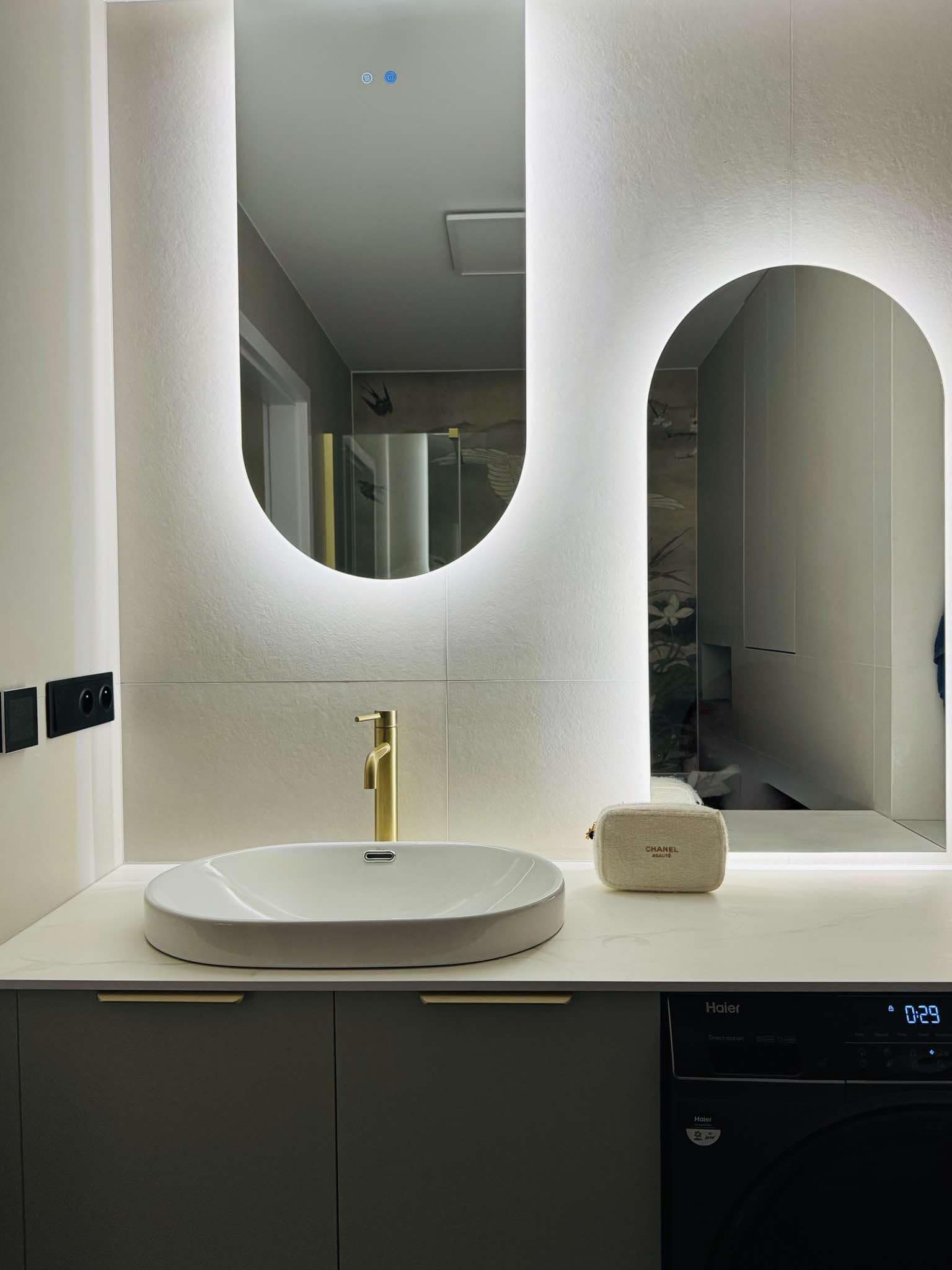 Nowoczesna łazienka z owalną umywalką, złotą baterią i podświetlanymi lustrami w kształcie łuków, z pralką Haier w zabudowie i kosmetyczką Chanel na blacie.