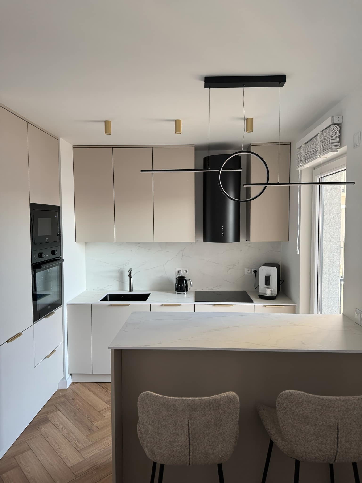 Jasna kuchnia w stylu minimalistycznym z drewnianą podłogą w jodełkę, czarnym okapem i nowoczesnym oświetleniem wiszącym z elementami w kształcie okręgu.