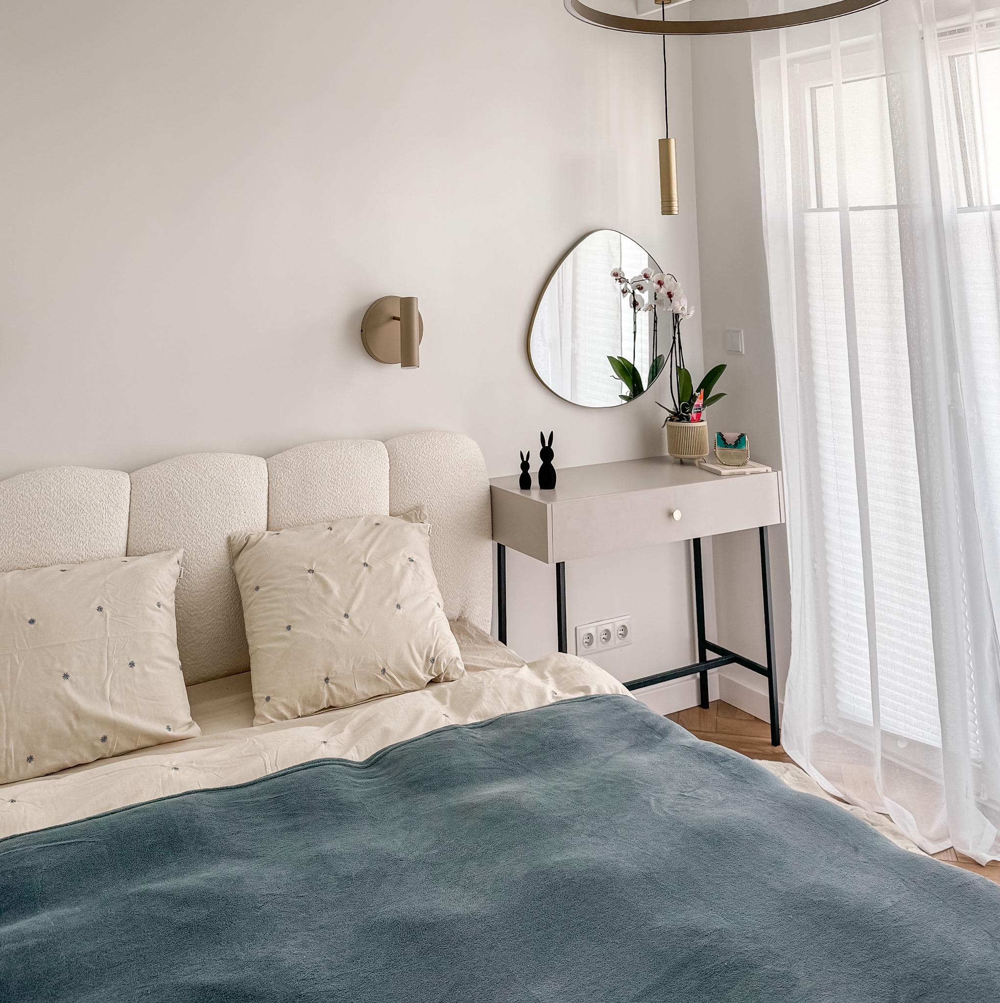 Jasna sypialnia z łóżkiem, toaletką z lustrem i dekoracjami. Minimalistyczny wystrój, naturalne światło, subtelne detale. Widok na okno z delikatnymi firankami.