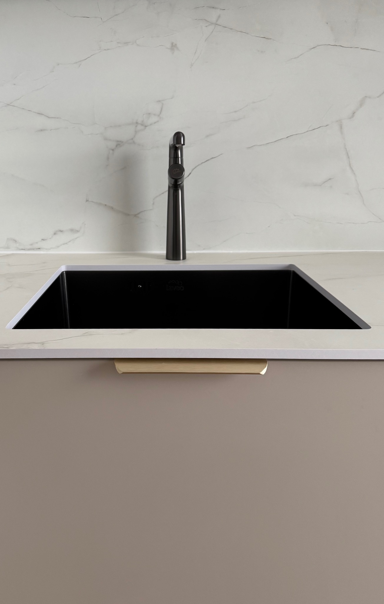 Czarne, prostokątne zlewozmywak w blacie z marmurowym wzorem, z ciemną baterią i beżową szafką z uchwytem. Minimalistyczny design, neutralna kolorystyka.