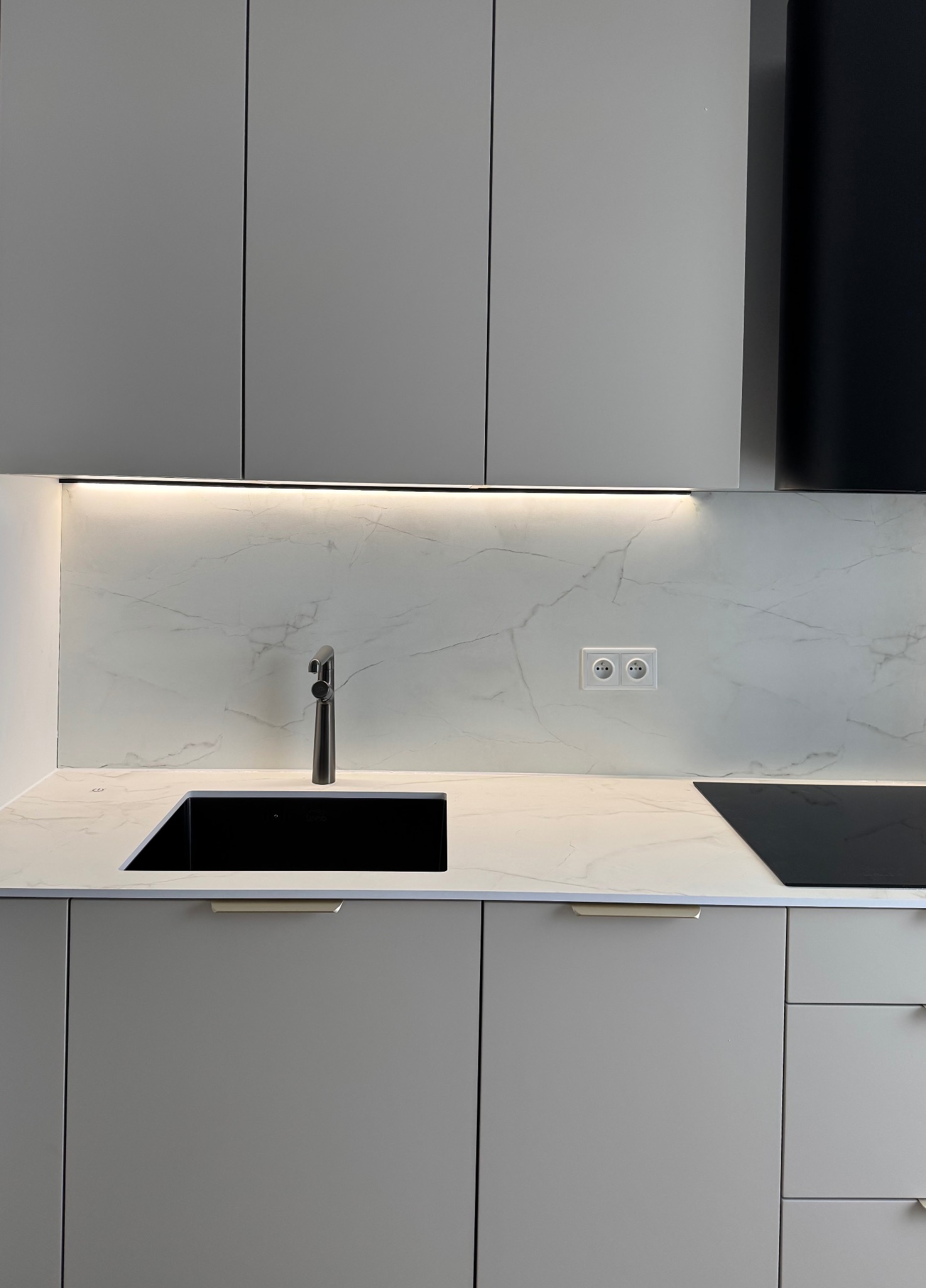 Minimalistyczna kuchnia: szare szafki, czarny zlew i bateria, marmurowy blat z podświetleniem LED. Gniazdka elektryczne na ścianie.