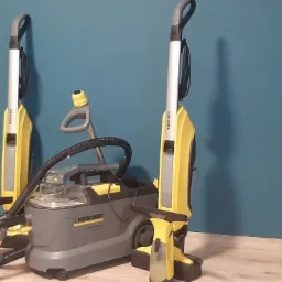 Dwa pionowe urządzenia Karcher do mycia okien i odkurzacz piorący Karcher na tle ściany w odcieniu petrolu i jasnej podłogi.