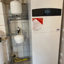 Instalacja pompy ciepła Ariston w Legnickim Polu, widoczne zbiorniki wyrównawcze, rury oraz panel sterowania, na ścianie naklejka informacyjna producenta.