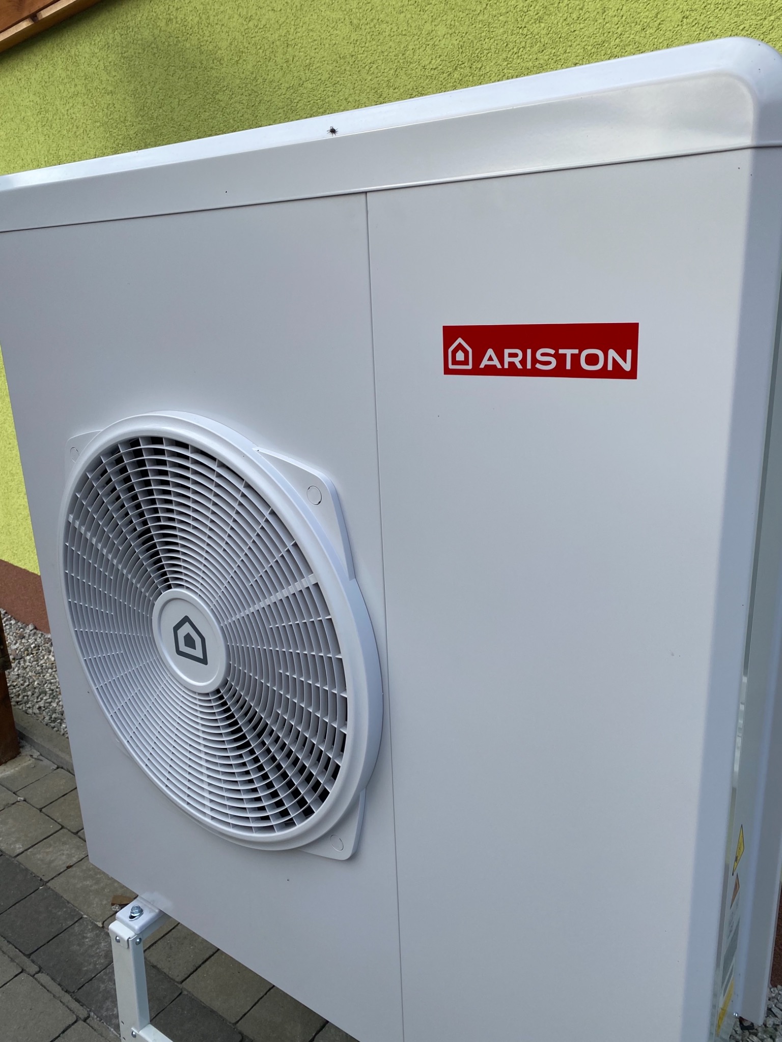 Biała pompa ciepła marki Ariston zamontowana na zewnątrz budynku z zieloną elewacją, widoczna kratka wentylatora i logo producenta.