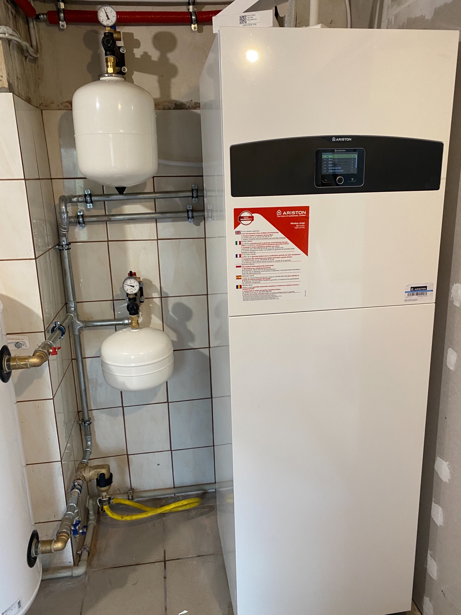 Instalacja pompy ciepła Ariston w Legnickim Polu, widoczne zbiorniki wyrównawcze, rury oraz panel sterowania, na ścianie naklejka informacyjna producenta.