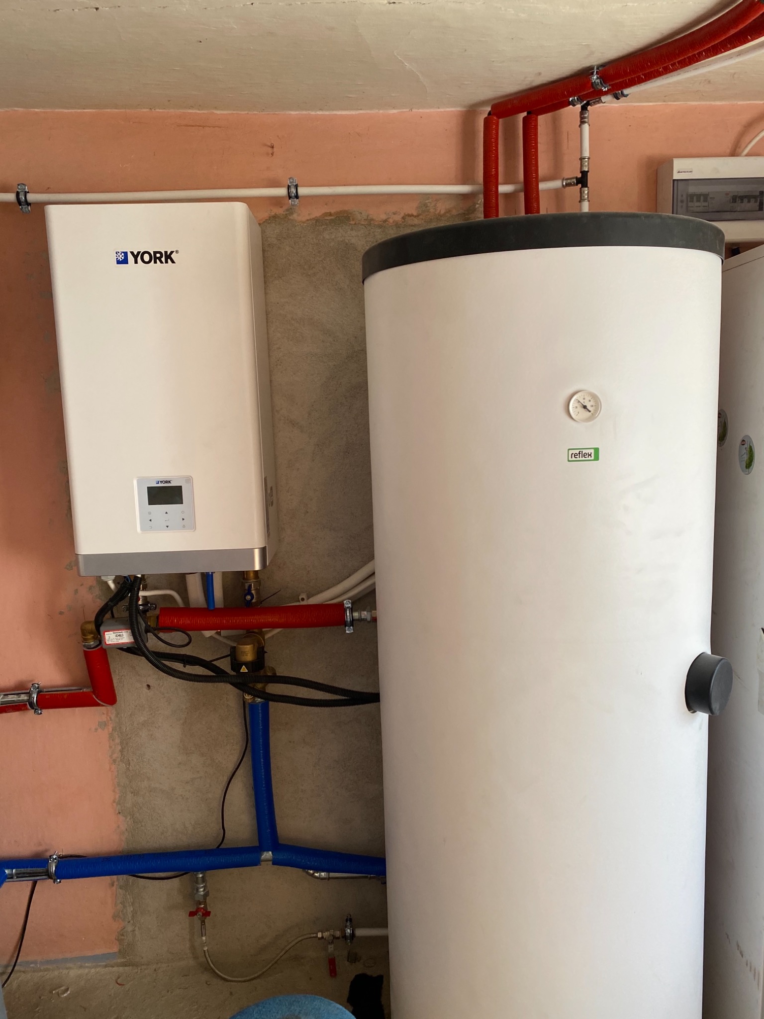 Biały zbiornik buforowy Reflex z termometrem i bojler York zamontowane na ścianie z widocznymi rurami instalacji grzewczej.