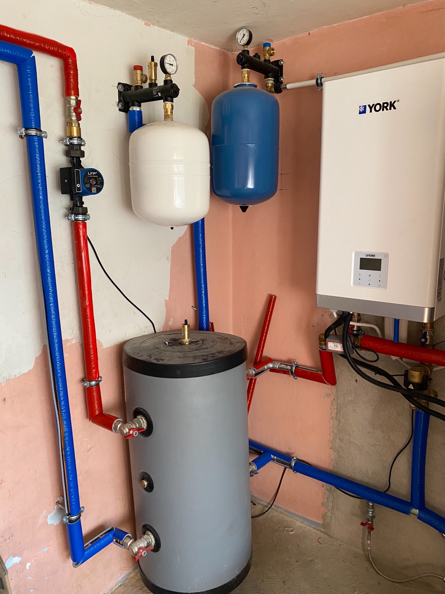 Instalacja grzewcza z szarym zasobnikiem ciepłej wody, biało-niebieskimi zbiornikami wyrównawczymi i białym kotłem York na tle surowych ścian.