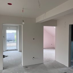 Dawid Rogalski - Wnętrze w trakcie prac wykończeniowych: świeżo pomalowane ściany na biało, surowa podłoga, widoczne instalacje elektryczne. W tle okno i różowe pomieszczenie.