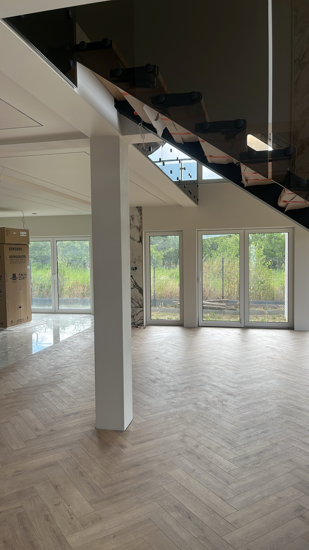 Jasne panele ułożone w jodełkę francuską w przestronnym wnętrzu z dużymi oknami i nowoczesnymi schodami ze szklaną balustradą. Widoczny fragment kolumny i karton lodówki.